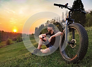 A man is sitting on the grass near a bicycle with thick wheels. Against the background of sunset
