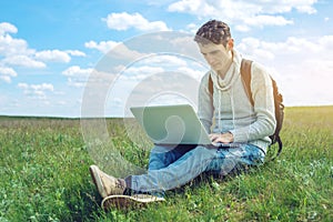 Man sitting on a green meadow with laptop wireless on the background of blue cloudy sky