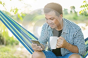 Man sitting on hammock and holding coffee cup and smartphone. Outdoor shooting with morning sunlight effect