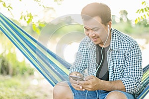 Man sitting on hammock and using smartphone with white earphone. Outdoor shooting with morning sunlight effect