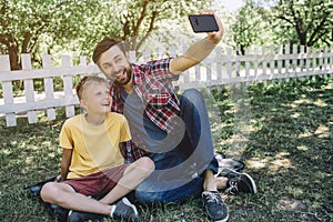 Man is sitting with his so on grass and holding phone in hand. He is taking selfie with kid. They are miling and posing