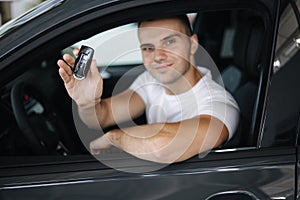 Man sitting in his new car in car showroom. Portrait of handsome man in car hold keys in hands