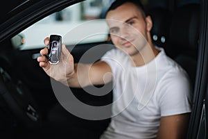 Man sitting in his new car in car showroom. Portrait of handsome man in car hold keys in hands. Keys in focus
