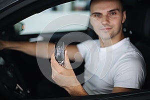 Man sitting in his new car in car showroom. Portrait of handsome man in car hold keys in hands. Keys in focus