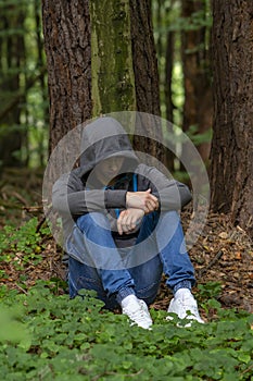 A man is sitting in a jacket with a hood hiding his face in the forest under a tree. Perhaps he is a lost tourist, a fugitive, an