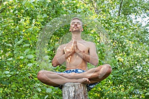 Man sitting and meditating with his hands together on trunk of tree on foliage background