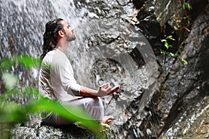 Man sitting in meditation yoga on rock at waterfall in tropical