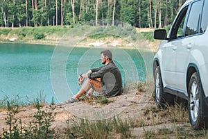 man sitting near white suv car at the edge looking at lake with blue water