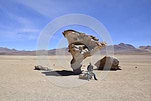 A man is sitting next to the famous stone tree rock formation (Arbol de Piedra) in the Siloli desert. Bolivia