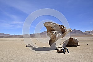 A man is sitting next to the famous stone tree rock formation (Arbol de Piedra) in the Siloli desert. Bolivia