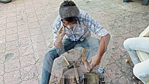 Man sitting outdoors using a mobile phone while working on a mechanical device with tools on the ground or electric welder on