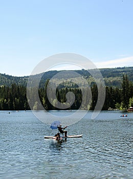 Man sitting and paddling under umbrella on mounting beach chair on top of SUP paddle board in the middle of Heffley Lake near Sun