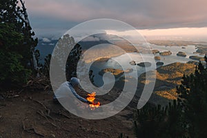 Man sitting on the rim with campfire above coastal view with islands and forests in cloudy weather from top view while sunset