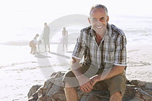 Man Sitting On Rock While Family Enjoying At Beach