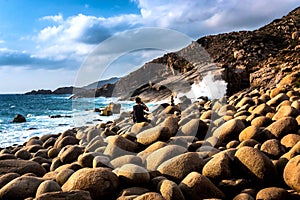 A Man Sitting on Rocks, Taking Photos of Waves at A Wild Eggshaped Rocks Beach with Dramatic Clouds in The Sky photo
