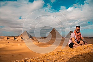 Man sitting on the sandy desert dunes posing in front of the great pyramids of giza. Traveling egypt in winter time, tourists