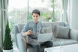 Man sitting on sofa with his laptop teleworking from home, wearing headphones and looking at the mobile phone