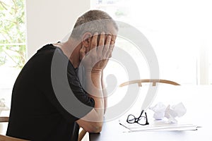 Man sitting at a table and hiding his head in despair. A pair of glasses lying on a pile of papers  with some crumpled papers on t