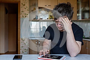 A man is sitting at a table at home. office at home. the man clutched his head, studying the information on the tablet, struggling