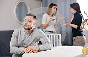 Man sitting at table while two women quarrel behind him