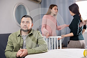 Man sitting at table while two women quarrel behind him