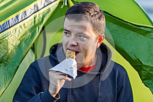 The man is sitting by the tent and eat snack