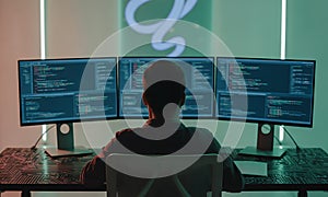 Man sitting at three monitors with code on screen from behind
