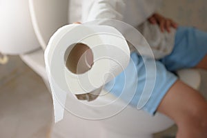 Man sitting on the toilet bowl and his hand is holding toilet paper or tissue close-up over blurred background in bathroom for