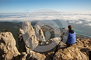 Man sitting with a tripod and photo camera on a high mountain peak above clouds, city and sea. Pro photographer