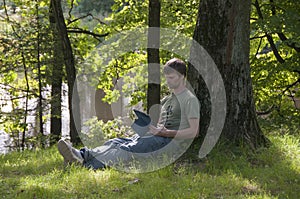 Man sitting under the tree by the lake and using his digital tablet. Guy working outdoors.