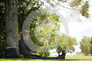 Man sitting under a tree, reposing and working on his computer. You can see his backpack next to him.