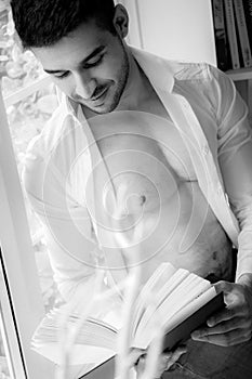 Man in sitting on window ledge with open shirt and pecs reading hardback book