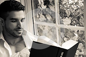 Man in sitting on window ledge with open shirt and pecs reading hardback book