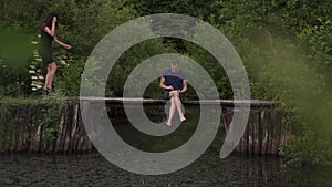 Man sitting on a wooden pontoon on a river in summertime and using a digital tablet while his wife passes deprived of