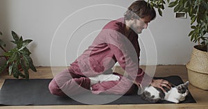 Man sitting on yoga mat cross legged stroking fluffy cat indoors natural light slow motion. Yogi master relaxing before