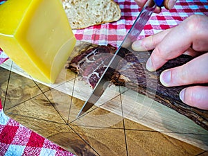 A man slicing smoked beef meat with his hands and holding a knife on a wooden cutting board with hard yellow cheese and bread on
