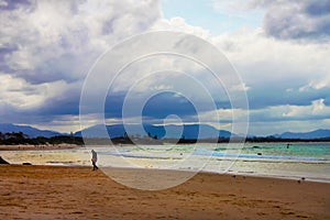 Man slumping - tired or sad - walking on beach coming out of surf at golden hour with beach full of people and mountians and storm photo