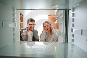 Man is smiling. Woman is complaining about an empty fridge to her husband on the kitchen