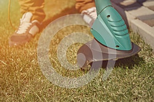 Man in sneakers and pants is mowing green grass with handheld electric lawn mower on his backyard. Gardening care