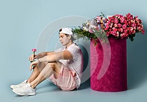 Man in sneakers shorts t-shirt is sitting at huge valentines day gift box with roses holding single flower in his hand