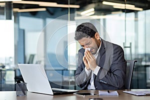 A man sneezes into a tissue at a workplace inside an office, a businessman is sick with a runny nose, works at a