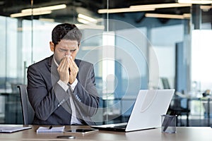 A man sneezes into a tissue at a workplace inside an office, a businessman is sick with a runny nose, works at a
