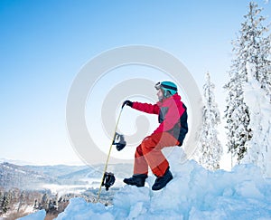 Man snowboarder sitting on top of a snowy hill with his snowboard enjoying stunning mountains view ski resort