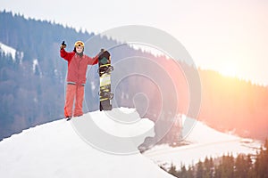 Man snowboarder standing on the top of the snowy slope with snowboard, showing thumbs up at winter ski resort