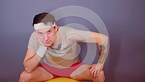 Man in sports headband jumping on gymnastic ball. Active male sits on fitness ball, propping his head on hand.