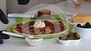 A man spreads mayonnaise on a base for a pizza smeared with tomato paste. Cooking ingredients are laid out nearby. Making pizza at