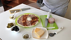 A man spreads mayonnaise on a base for a pizza smeared with tomato paste. Cooking ingredients are laid out nearby. Making pizza at