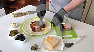 A man spreads tomato paste on a pizza base. Cooking ingredients are laid out nearby. Making pizza at home