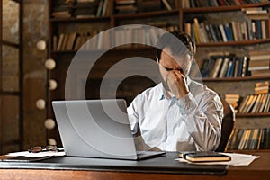 A man squints eyes and covers face with hand tiredly, sits at a table in the library and uses a laptop. A respectable photo