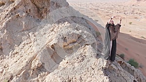 A man with a staff in his hand and the clothes of a shaman stands on a rock among the sands of the desert and dances